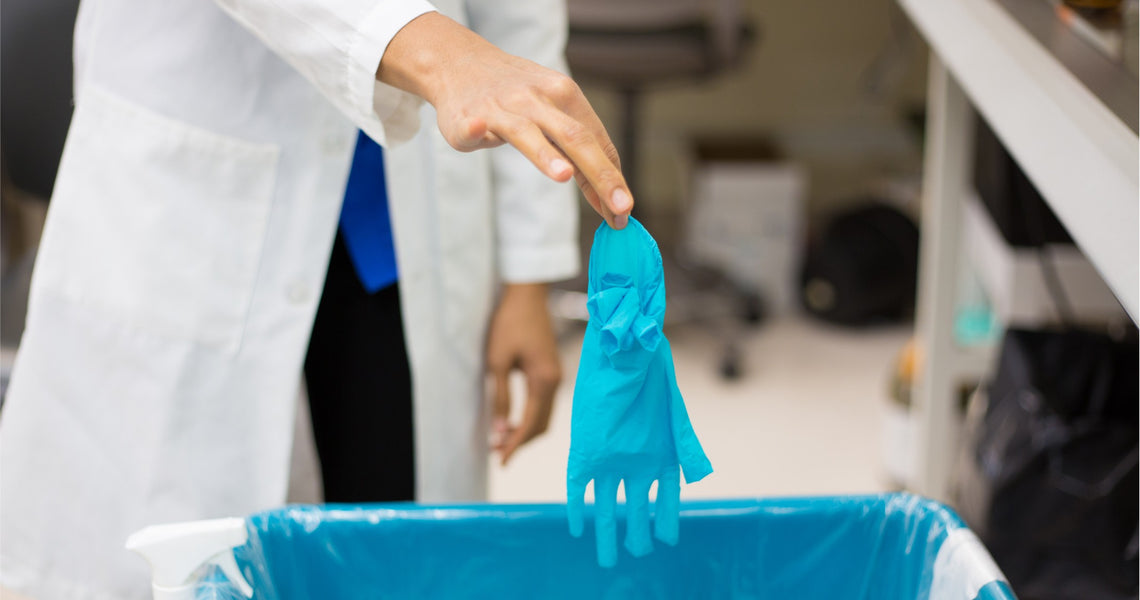 A person in a lab coat disposes of a blue glove in a waste bin.