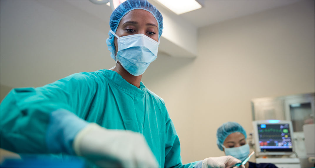 A surgeon wearing a mask, gloves, and surgical scrubs performs an operation in an operating room, with a medical monitor visible in the background.