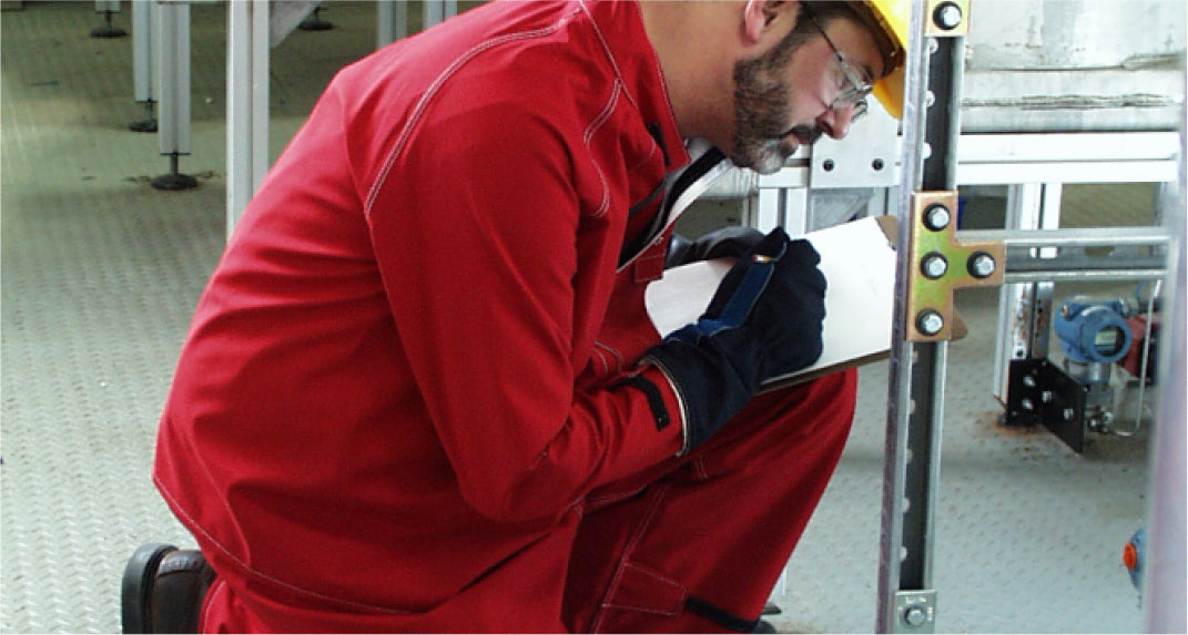 A worker wearing a red jumpsuit and yellow hard hat kneels on the ground while writing on a clipboard in an industrial setting.