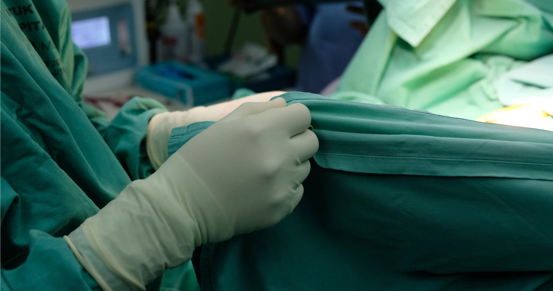 A healthcare professional in green scrubs and gloves holds a sterile drape in a hospital setting.