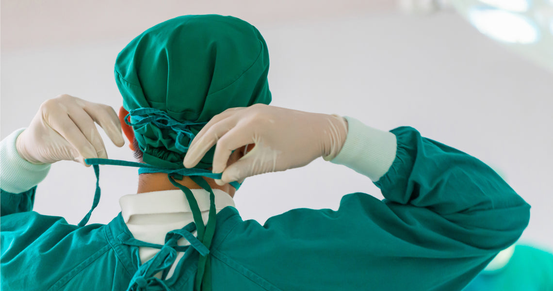 A surgeon in green scrubs and gloves ties the strings of their surgical mask, preparing for surgery.