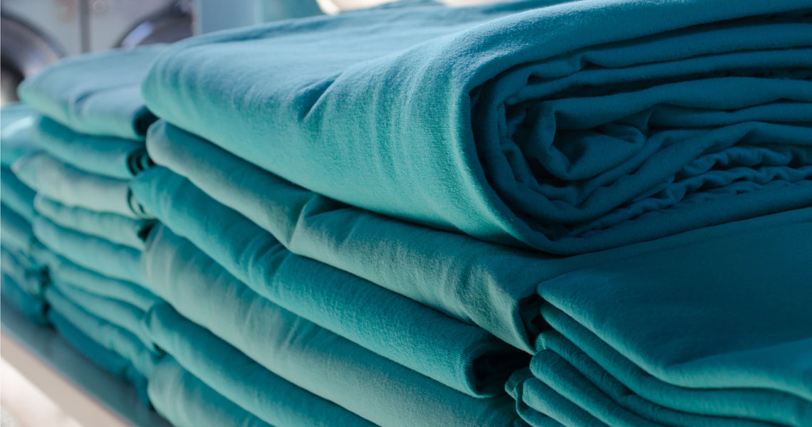 Stacks of neatly folded teal-colored sheets are arranged on a shelf, with light highlighting their smooth texture.