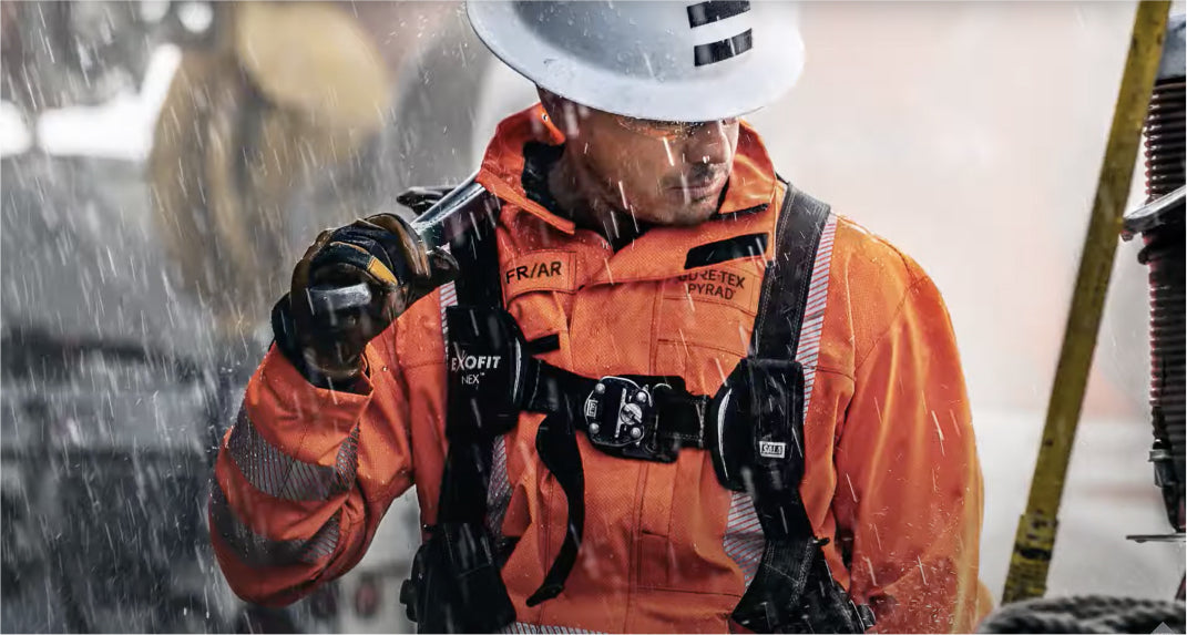 A worker dressed in an orange safety jacket and white helmet stands in the rain, adjusting his gear with a focused expression.