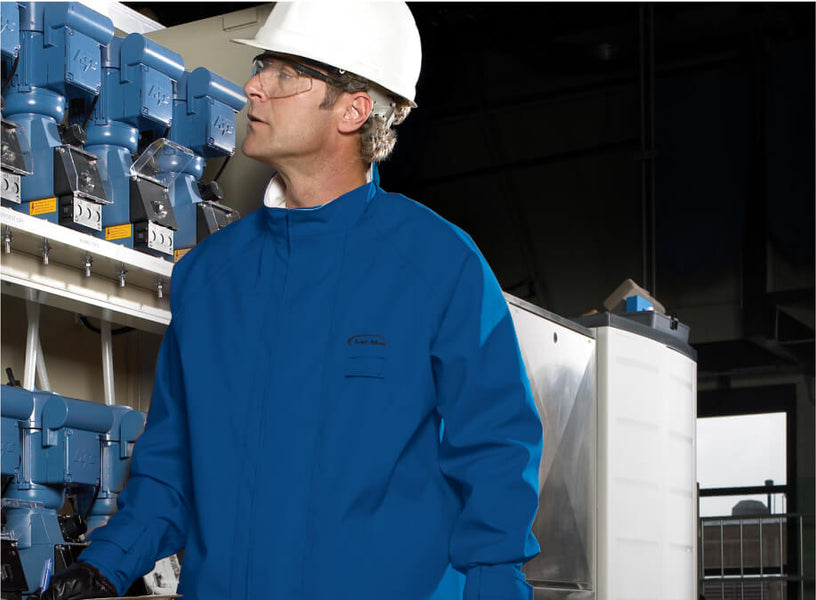 A worker wearing a blue protective suit, safety glasses, and a white hard hat stands near industrial equipment, looking towards the left side of the frame.