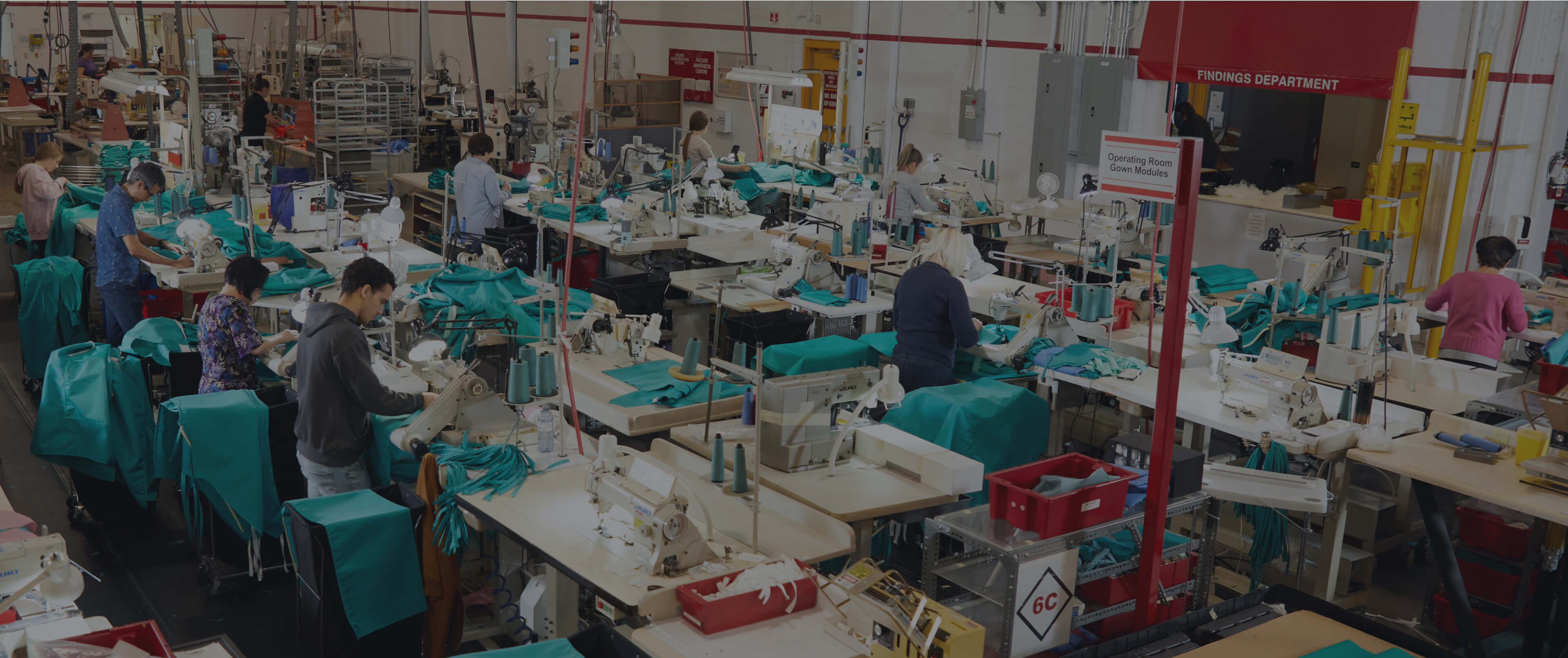 A busy factory of Lac Mac with workers at sewing machines and tables, producing green garments. Various materials and equipment are organized on workstations throughout the space.